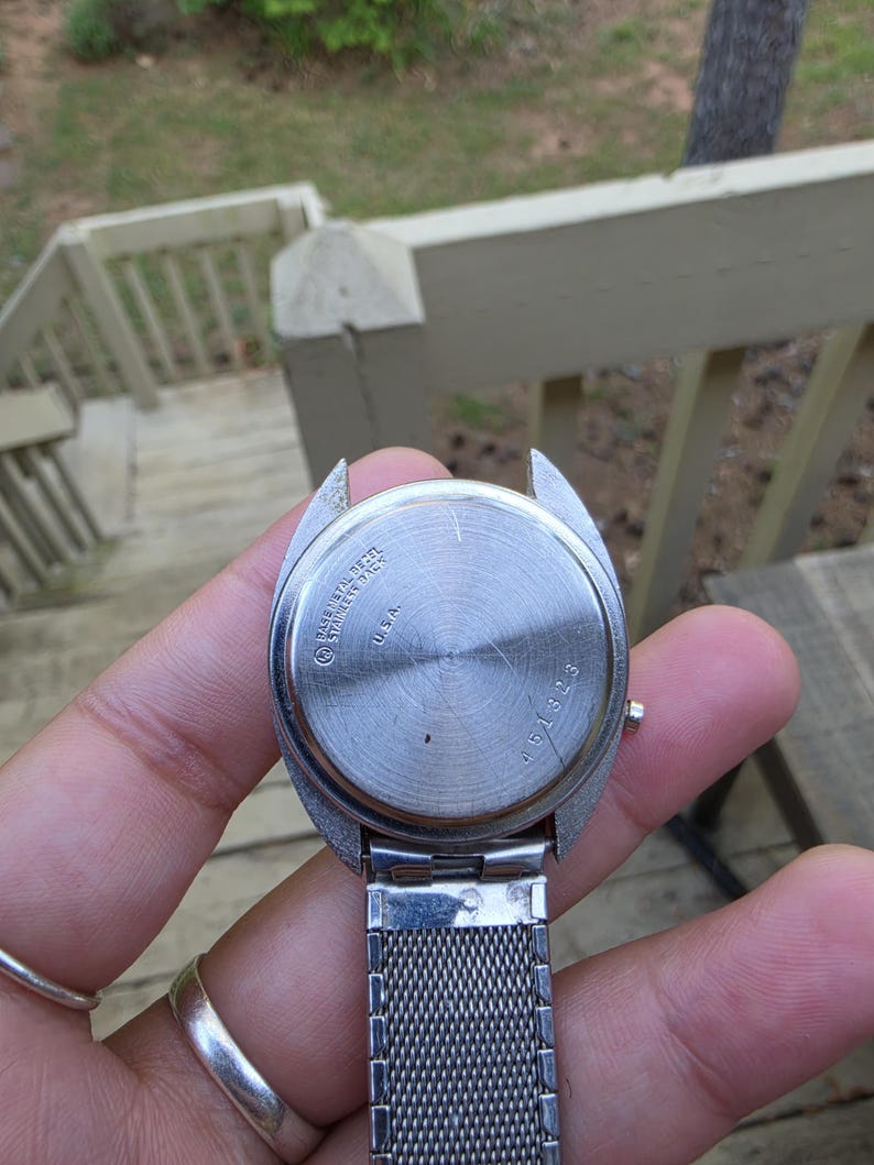 May include: A stainless steel watch with a silver mesh band. The watch face is silver with the text "BASE METAL STAINLESS STEEL BACK U.S.A." and the number "451323". The watch is held in a hand.