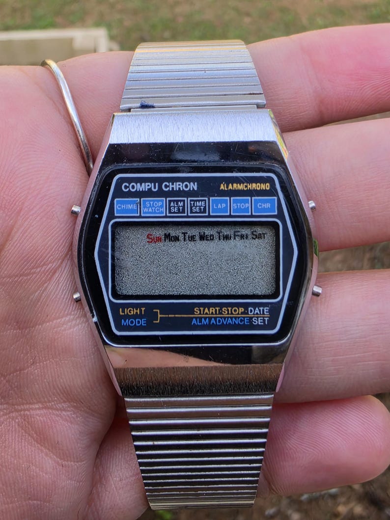 Może przedstawiać: Zegarek cyfrowy Compu Chron w stylu vintage ze srebrnym metalowym paskiem. Czarna tarcza zegarka wyświetla dzień tygodnia i r&oacute;żne przyciski funkcyjne. Zegarek ma napis "COMPU CHRON" i "ALARMCHRONO".