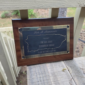 Puede incluir: Placa de madera con una placa de metal oscuro con un mapa grabado de Puerto Rico y texto. El texto incluye "FBI SAN JUAN TERRORISM SQUAD" y fechas. La placa tiene un borde decorativo.