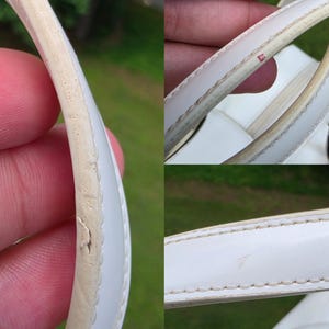 Op de afbeelding: Een wit handtashengsel met zichtbare slijtage langs de randen. Het hengsel vertoont tekenen van barsten, afbladdering en verkleuring op de rand, samen met een kleine rode vlek en lichte krassen op het witte materiaal.
