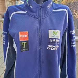 May include: A blue Yamaha zip-up jacket with white accents and multiple sponsor logos, including Movistar, Eneos, and Monster Energy. The jacket has a high collar and a full-length zipper. The sleeves are dark blue with white stripes.
