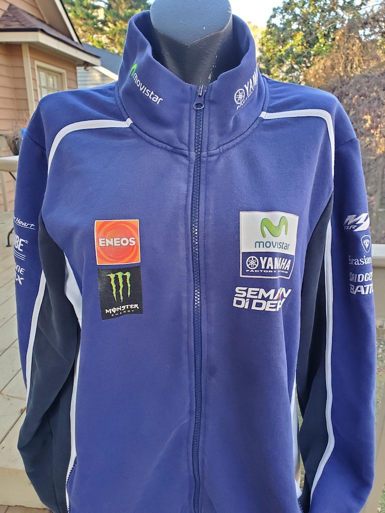 May include: A blue Yamaha zip-up jacket with white accents and multiple sponsor logos, including Movistar, Eneos, and Monster Energy. The jacket has a high collar and a full-length zipper. The sleeves are dark blue with white stripes.
