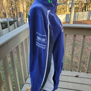 May include: A blue zip-up jacket with white and black accents. The jacket features the Movistar logo in green on the collar and sponsor logos on the sleeve, including "Yamalube" and "Bridgestone". The jacket is displayed on a stand outdoors.
