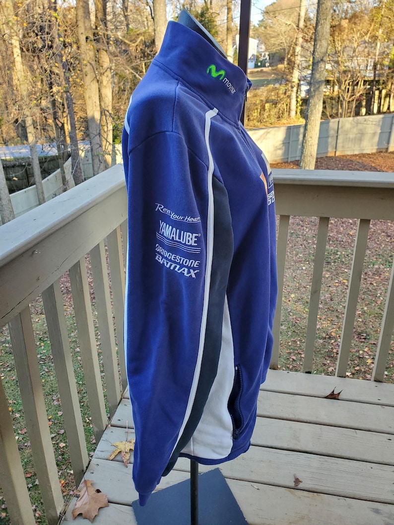 May include: A blue zip-up jacket with white and black accents. The jacket features the Movistar logo in green on the collar and sponsor logos on the sleeve, including "Yamalube" and "Bridgestone". The jacket is displayed on a stand outdoors.