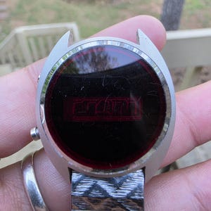 May include: Vintage silver-toned digital watch with a rectangular red display. The watch face is round with a dark red screen displaying the time. The watch band is a silver-toned, patterned metal.