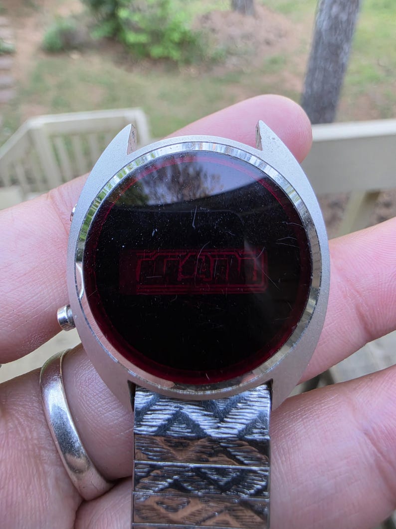 May include: Vintage silver-toned digital watch with a rectangular red display. The watch face is round with a dark red screen displaying the time. The watch band is a silver-toned, patterned metal.