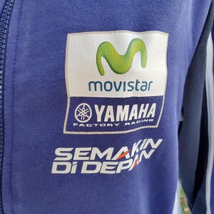 May include: A blue zip-up jacket featuring the Movistar and Yamaha Factory Racing logos. The jacket has white and green accents, with the text "SEMAKIN DI DEPAN" printed in white and orange.