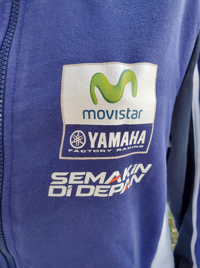May include: A blue zip-up jacket featuring the Movistar and Yamaha Factory Racing logos. The jacket has white and green accents, with the text "SEMAKIN DI DEPAN" printed in white and orange.