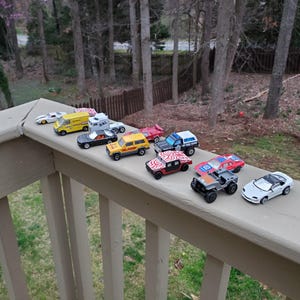 May include: A collection of toy cars, including a yellow delivery truck, a black car, a yellow SUV, a black and white SUV, a red sports car, a green jeep, and a white sports car. The cars are lined up on a wooden deck railing.