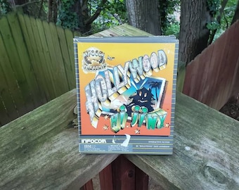 Può includere: Una scatola di gioco vintage per IBM PC. La scatola è gialla e nera con il testo "Infocom Presents Hollywood Hijinx" e un'immagine di cartone animato di un cartello di Hollywood con un'auto che lo attraversa.