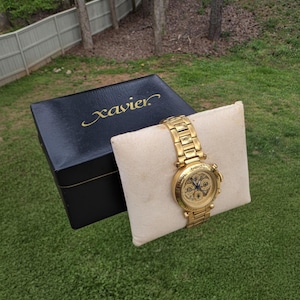 May include: A gold-toned wristwatch with a matching band rests on a cream-colored pillow. The watch face features multiple subdials. A black box with the name "Xavier" in gold lettering sits nearby. The watch and box are on a green grassy surface.
