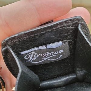 May include: Black leather coin purse with a textured strap. The interior features a tag with the brand name "Brighton" in a cursive font. The purse is open, revealing the inside. The strap is a dark brown color.