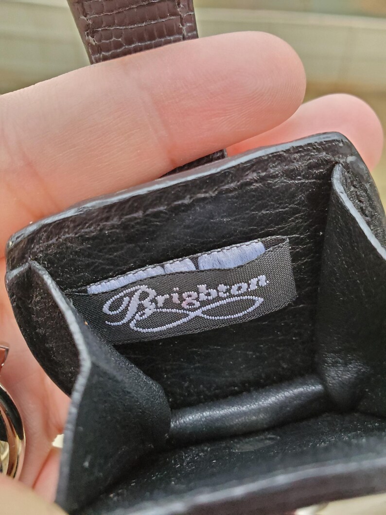 May include: Black leather coin purse with a textured strap. The interior features a tag with the brand name "Brighton" in a cursive font. The purse is open, revealing the inside. The strap is a dark brown color.