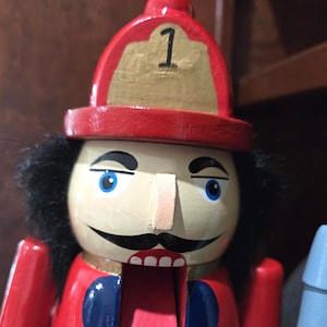 May include: A wooden nutcracker in the shape of a firefighter. The nutcracker is painted red with a gold-colored helmet and the number 1. It has black hair, blue eyes, and a black mustache. The mouth is open, revealing white teeth and a pink interior.