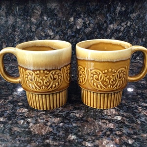 May include: Two brown ceramic mugs with a decorative floral pattern. The mugs have a light brown glaze and a darker brown band around the bottom.