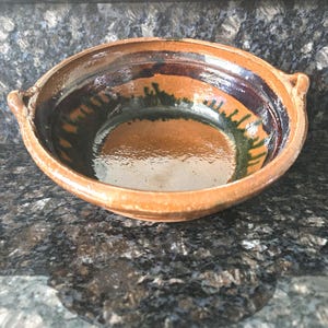 May include: A handmade, round, earthenware bowl with a wide rim and two small handles. The bowl features a brown exterior and interior with dark brown, green, and black accents. The bowl is likely used for serving food.