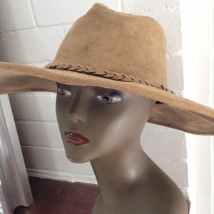 May include: A tan suede-like hat with a wide brim and a braided band around the crown. The hat is displayed on a mannequin head. The hat's brim is wide and slightly curved, and the crown is a soft, rounded shape.