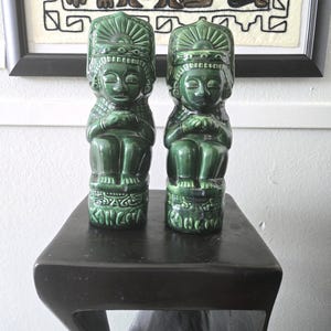 May include: A white ceramic mug with a black handle and the text "But First, Coffee" in a bold, black font. The mug is set against a blurred background, suggesting a cozy setting.