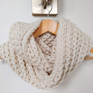 May include: Cream-colored, chunky knit infinity scarf displayed on a wooden hanger. The scarf is made of thick yarn, showcasing a textured, ribbed pattern. The neutral color and cozy design suggest warmth and comfort, suitable for cold weather.