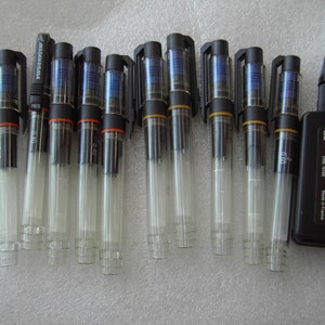 Może przedstawiać: Zestaw dziesięciu czarnych i przezroczystych,  napełnianych technicznych długopisów z czarnym flakonem z atramentem. Długopisy mają końcówkę o grubości 0,40 mm i są oznaczone "Mecanorama".