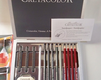 Può includere: Un set di matite da disegno Cretacolor in una custodia argentata. Il set include una varietà di matite a grafite, una matita bianca, una gomma da cancellare impastabile, un temperamatite e un blocco di levigatura. La custodia è etichettata "Cretacolor, Vienna. Una passione per la matita."