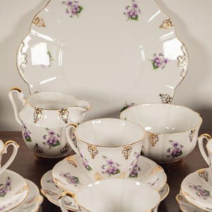May include: A vintage tea set featuring white porcelain cups, saucers, and a serving plate. Each item is adorned with purple floral designs and gold edging. Includes a creamer and sugar bowl.