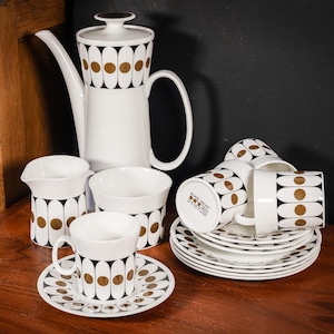 Może przedstawiać: Biały ceramiczny zestaw do herbaty z geometrycznym wzorem w kolorze czarnym i złotym. Zestaw zawiera czajnik, dzbanek na śmietanę, cukiernicę i cztery filiżanki i spodki.