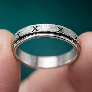 May include: A silver ring with a textured band and a series of engraved 'X' marks. The ring is held between two fingers against a blurred teal background. The ring's design includes a spinning center band.