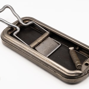 May include: A rectangular, metallic, vintage-style tobacco tin or cigarette case with a hinged, spring-loaded lid mechanism. The exterior has a muted, brushed silver finish. Inside, a metal plate is engraved with the text 'Tony Royer' and 'Paris'.
