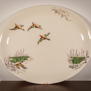 May include: A large, round, off-white ceramic plate with a painted duck scene. The plate features several flying ducks in shades of brown, orange, and green, above a marsh scene with reeds and water. The plate is approximately 12 inches in diameter.