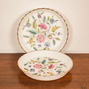 May include: Two white ceramic plates with a floral design. The plates have a scalloped gold rim and feature pink, blue, yellow, and green flowers and leaves. The plates are on a wooden surface.