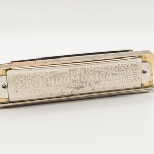 May include: A vintage Hohner Super Chromonica harmonica. The instrument has a silver body with gold accents. The text "Super Chromonica" and "M. Hohner" are visible. The harmonica has a small silver mouthpiece. The instrument is approximately 10 cm long.