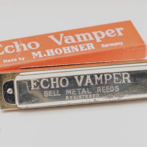 May include: A vintage Echo Vamper harmonica with a silver finish and gold-toned metal accents, next to its orange box. The box is labelled "Echo Vamper" and "M. Hohner Germany".