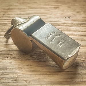 May include: A vintage metal whistle with a key ring, resting on a wooden surface. The whistle is engraved with "The Acme Thunderer" and "Made in England". The whistle has a rectangular body and a rounded mouthpiece.