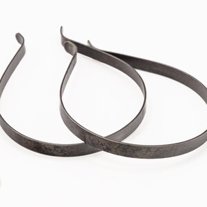 May include: Two dark grey metal headbands with a weathered, matte finish. Each headband features a simple, thin, flexible band design with slightly curved ends. The metal surface shows a textured, aged appearance with subtle variations in colour.