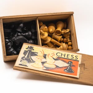 Op de afbeelding: Houten schaakset met zwarte en natuurlijk gekleurde stukken in een verdeelde houten doos. De doos heeft een schuifdeksel met het woord "CHESS" en illustraties van schaakstukken.
