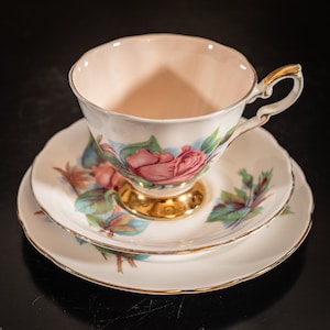 Könnte beinhalten: Ein weißes Tee-Set mit einem floralen Muster aus rosa Rosen. Die Teetasse hat ein rosafarbenes Inneres und einen goldenen Rand. Die Untertasse hat einen goldenen Rand und eine kleinere Untertasse, die in sie eingebettet ist.