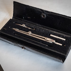 Puede incluir: Un juego de compás plateado con una caja de cuero negro. El compás tiene un brazo de extensión de metal y un accesorio de lápiz.