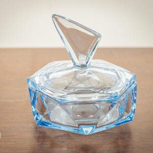 Op de afbeelding: Een zeshoekige, lichtblauwe glazen sieradendoos met een heldere, driehoekige handgreep op het deksel. De doos wordt gepresenteerd op een houten oppervlak, waarbij het geometrische ontwerp en de kleur worden getoond.