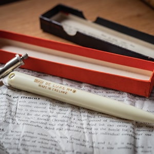 May include: A white stencil pen with gold lettering that reads "DE LUXE NO. 9 MADE IN ENGLAND". The pen is in a red and black box. The pen is on a piece of paper with text about stencil care.