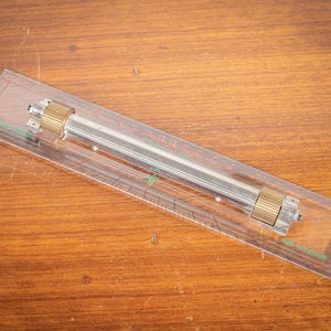 Puede incluir: Una herramienta de dibujo de plástico transparente y metal con una regla y un diseño cilíndrico. La herramienta tiene una barra central plateada con extremos cilíndricos dorados. La herramienta está sobre una superficie de madera.