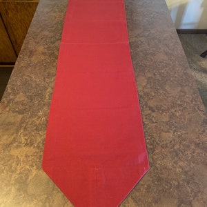 Puede incluir: Un camino de mesa de color burdeos con un extremo puntiagudo. El camino de mesa está hecho de una tela lisa y tiene un brillo sutil.