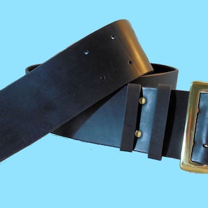 May include: A wide black leather belt with a brass buckle. The belt is folded in half and the buckle is on the right side of the image.