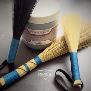May include: Two small handmade brooms with blue wrapped handles and dark bristles, next to a jar of turquoise glaze. The jar is labeled "Spectrum Raku Glaze" and "852 Turquoise". The brooms have black loops attached to the handles. The text "HADENMADE" is also visible.