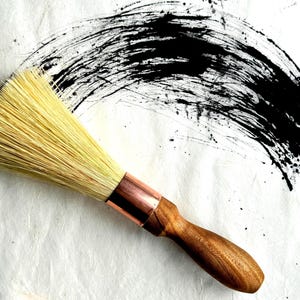 May include: A round brush with light-colored natural bristles, a polished copper ferrule, and a smooth, contoured wooden handle. The brush is positioned diagonally across a white surface featuring a wide, textured black ink brushstroke.