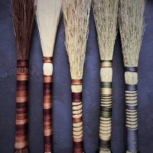 May include: Five decorative brooms with natural bristles and intricately wrapped handles. The handles are wrapped with twine in shades of brown, beige, and green. These handcrafted brooms are ideal for display or as a unique gift item.