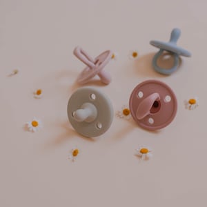 May include: Four pastel-colored pacifiers in shades of pink, blue, and green are scattered on a light pink surface, interspersed with small white and yellow flowers. The pacifiers have a rounded shield and a nipple-shaped teat.