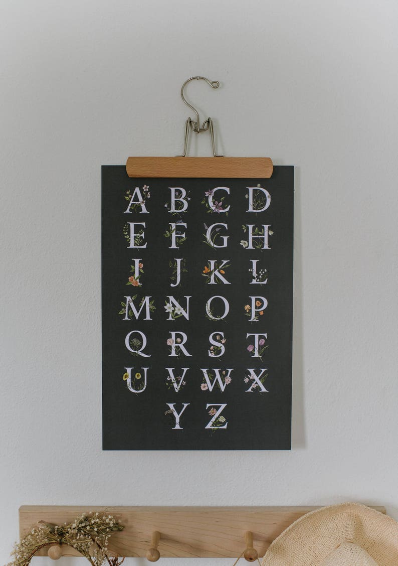 May include: A black and white alphabet poster with floral accents. Each letter is surrounded by a variety of flowers and greenery. The poster is hanging on a white wall with a wooden clip.