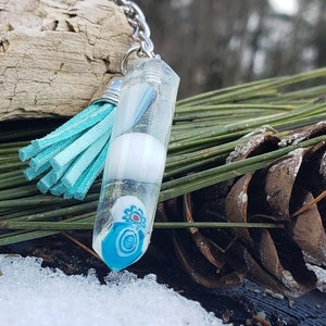 Op de afbeelding: Een transparante glazen hanger met een blauw en wit swirl-ontwerp en een witte kraal erin. De hanger is bevestigd aan een zilveren ketting met een turquoise kwast.