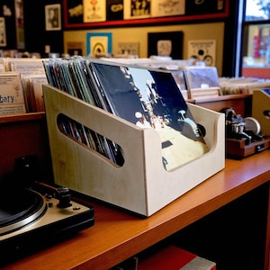 Può includere: Una cassa di legno chiaro piena di dischi in vinile. La cassa, con maniglie ritagliate, è appoggiata su una mensola di legno. Un giradischi è in primo piano, con altri dischi visibili sullo sfondo.
